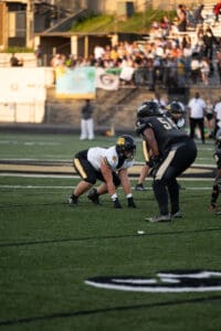 High school football game with players in action on the field, spectators in the background.