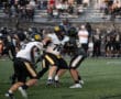 High school football players competing in a game, with spectators in the stands during a sunny afternoon.