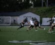 High school football game action, players in black and white jerseys on the field during a play at dusk.