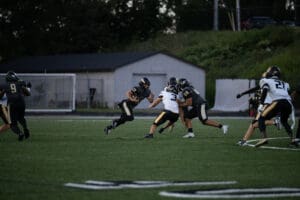 High school football game action, players in black and white jerseys on the field during a play at dusk.