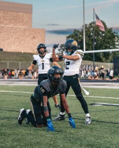 Football players on the field, with one kneeling and two celebrating, during a game with American flag in the background.