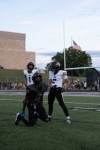 Football players on field during a game, with one kneeling, American flag in background.