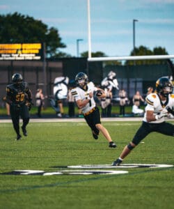 High school football quarterback runs with the ball during a game, evading a defender.