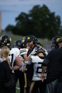 Football coach giving instructions to players on the sidelines during a game at dusk.
