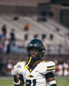 Football player smiling on field during a game, wearing black helmet and white jersey number 21.