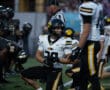 Football players on field during night game, teammates in action, focused expressions, wearing black and yellow uniforms.