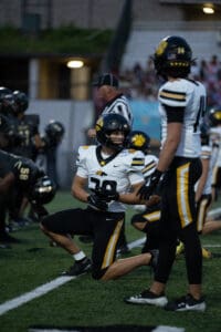 Football players on field during night game, teammates in action, focused expressions, wearing black and yellow uniforms.