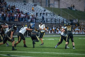High school football game action with quarterback preparing to throw the ball, players in motion on the field.