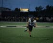 Football player running with the ball during a night game on a field, with blurred crowd in the background.