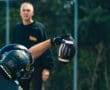 Football player in black helmet catching a football during practice, coach observing in the background.