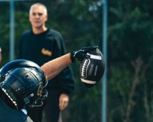 Football player in black helmet catching a football during practice, coach observing in the background.
