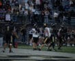 High school football game action, players on field, cheering crowd in bleachers, night scene.