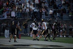 High school football game action, players on field, cheering crowd in bleachers, night scene.