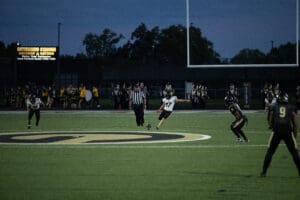 Football kickoff at Antimarino Stadium, players and referee in action, marching band and crowd in the background.