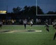Football kickoff at nighttime with players on the field, referee in striped shirt, and band in the background.