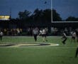 High school football players during a night game with a ball in the air, referee and band visible in background.