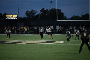 High school football players during a night game with a ball in the air, referee and band visible in background.
