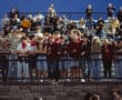 Crowd in bleachers at outdoor event, dressed in casual and cowboy attire, under clear blue sky.