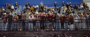 Crowd in bleachers at outdoor event, dressed in casual and cowboy attire, under clear blue sky.