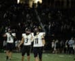 Football players on the field during a night game, wearing black and white uniforms, with a referee nearby.