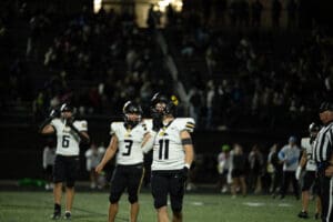 Football players on the field during a night game, wearing black and white uniforms, with a referee nearby.