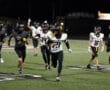 Football player sprints downfield with the ball during nighttime game, teammates in pursuit on a lit field.