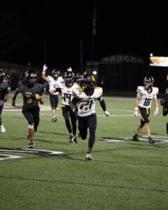 Football player sprints downfield with the ball during nighttime game, teammates in pursuit on a lit field.