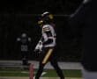 Football player in black and yellow uniform walking on field at night with helmet in hand.