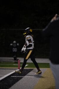 Football player in black and yellow uniform walking on field at night with helmet in hand.