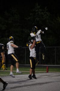 Football players celebrate a touchdown with a jump and lift under stadium lights during a night game.