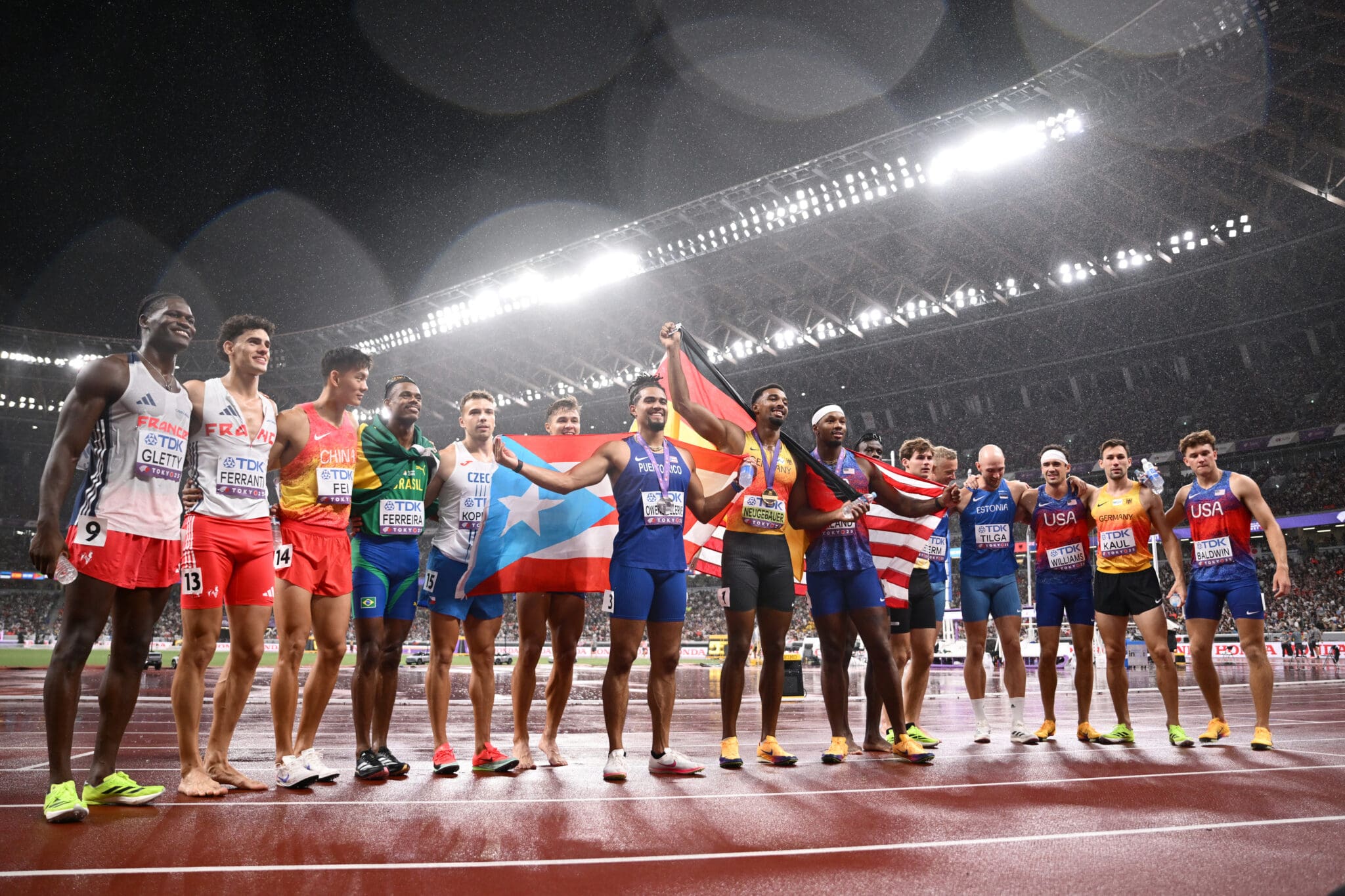 Athletes from various countries celebrate on a wet track with flags at an international sports event.