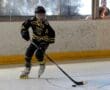 Youth inline hockey player in action on the rink, wearing black and yellow gear, with an audience watching through glass.