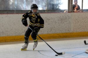 Youth inline hockey player in action on the rink, wearing black and yellow gear, with an audience watching through glass.