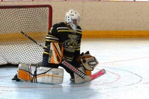 Roller hockey goalie in action, wearing protective gear, kneeling in front of net on rink.