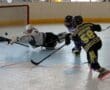 Roller hockey action shot with player attempting a goal, goalie making a diving save in an indoor rink.