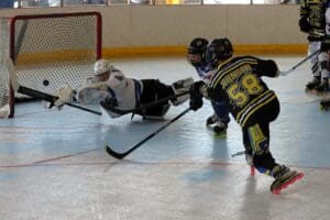 Roller hockey action shot with player attempting a goal, goalie making a diving save in an indoor rink.
