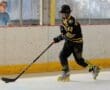Roller hockey player in action with black and yellow gear on rink, coach observing in the background.