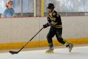 Roller hockey player in action with black and yellow gear on rink, coach observing in the background.