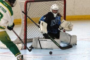 Roller hockey goalie defends against incoming puck, wearing blue jersey and white gear, focused on blocking the shot.