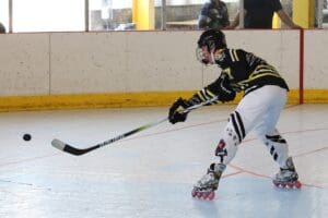 Roller hockey player in action on the rink, taking a shot with a black stick, wearing protective gear and skates.