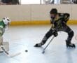 Roller hockey player in black jersey aiming puck at goalie in green jersey on indoor rink.