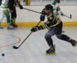 Roller hockey player in black and yellow shooting puck during intense game on indoor rink.