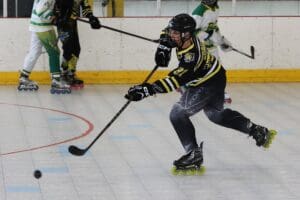 Roller hockey player in black and yellow shooting puck during intense game on indoor rink.