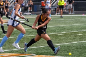 Field hockey players in action, one wearing a black uniform hitting a yellow ball on a green field during a match.