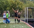 Field hockey players celebrating a goal on a sunny day, with teammates and a goalie nearby on the field.