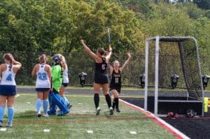 Field hockey players celebrating a goal on a sunny day, with teammates and a goalie nearby on the field.