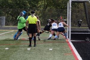 Field hockey players competing near goal, with referee and goalkeeper, on a sunny day. Game action close-up.