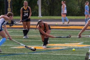 Field hockey game in action, player in black uniform strikes the ball on a green turf.