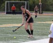 Female field hockey player in action on a turf field, wearing a black Tigers uniform with goalie and teammates nearby.