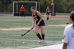 Female field hockey player in action on a turf field, wearing a black Tigers uniform with goalie and teammates nearby.